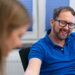 Mann im blauen Poloshirt sitzt am Tisch, im Vordergrund ist eine Person unscharf.