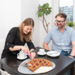 Frau schneidet Kuchen an einem Tisch, Mann sitzt daneben.